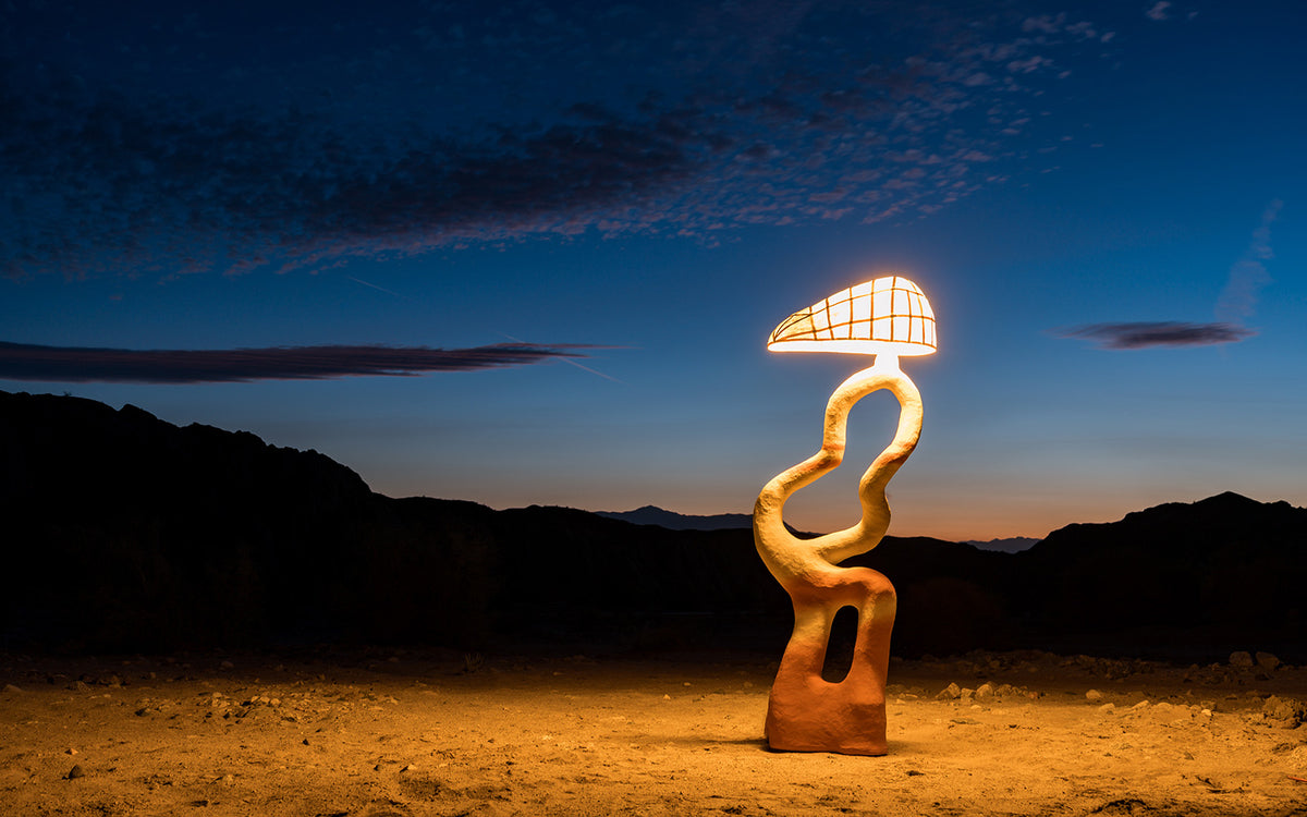 A sculptural biomorphic floor lamp stands on a sandy desert floor at dusk, with dark mountain silhouettes and a deep blue sky in the background. The mulberry paper shade glows with a warm yellow light, and the base is a textured terracotta and sand color.