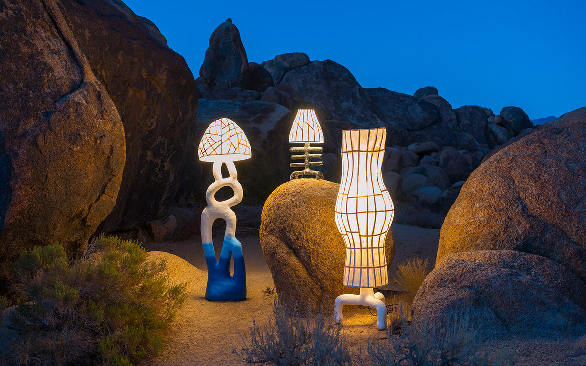 Three sculptural lamps in a desert rocky setting at dusk, glowing with warm light against blue sky and tan boulders.
