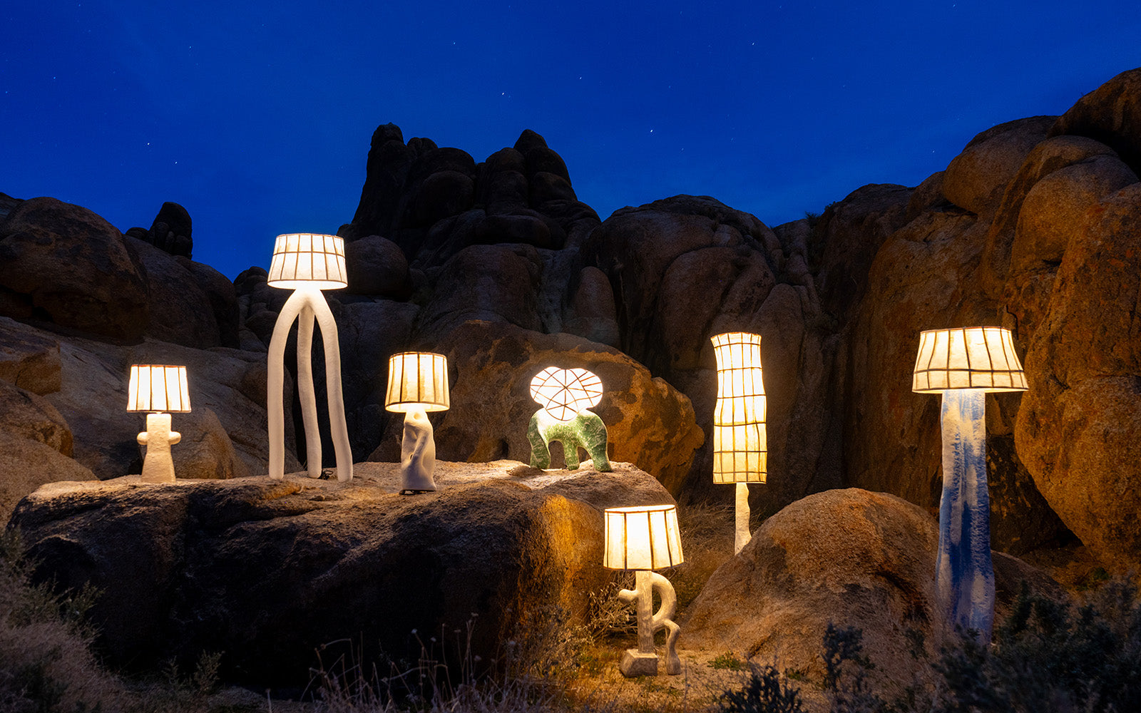 A collection of whimsical, sculptural table and floor lamps with mulberry paper shades and organic, abstract bases displayed outdoors on large desert rock formations under a dark blue night sky.