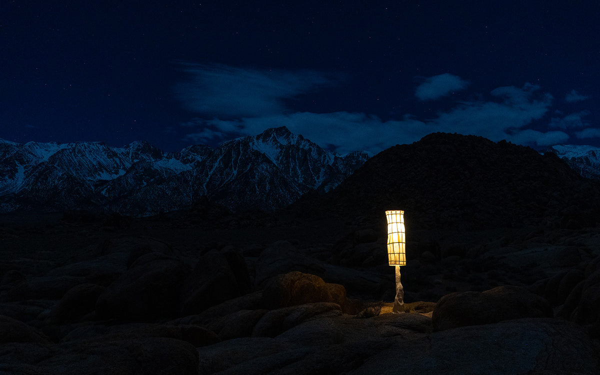 A 7' tall, glowing lantern-style lamp stands on rocky ground at night, with the dark snow capped sierra Nevada mountains  and a starry sky in the background.