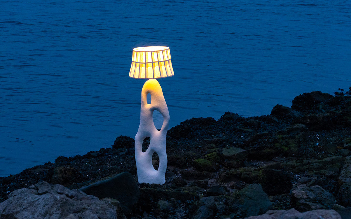 A sculptural white floor lamp with a mulberry paper shade stands lit on a rocky shoreline at dusk, with deep blue water in the background.