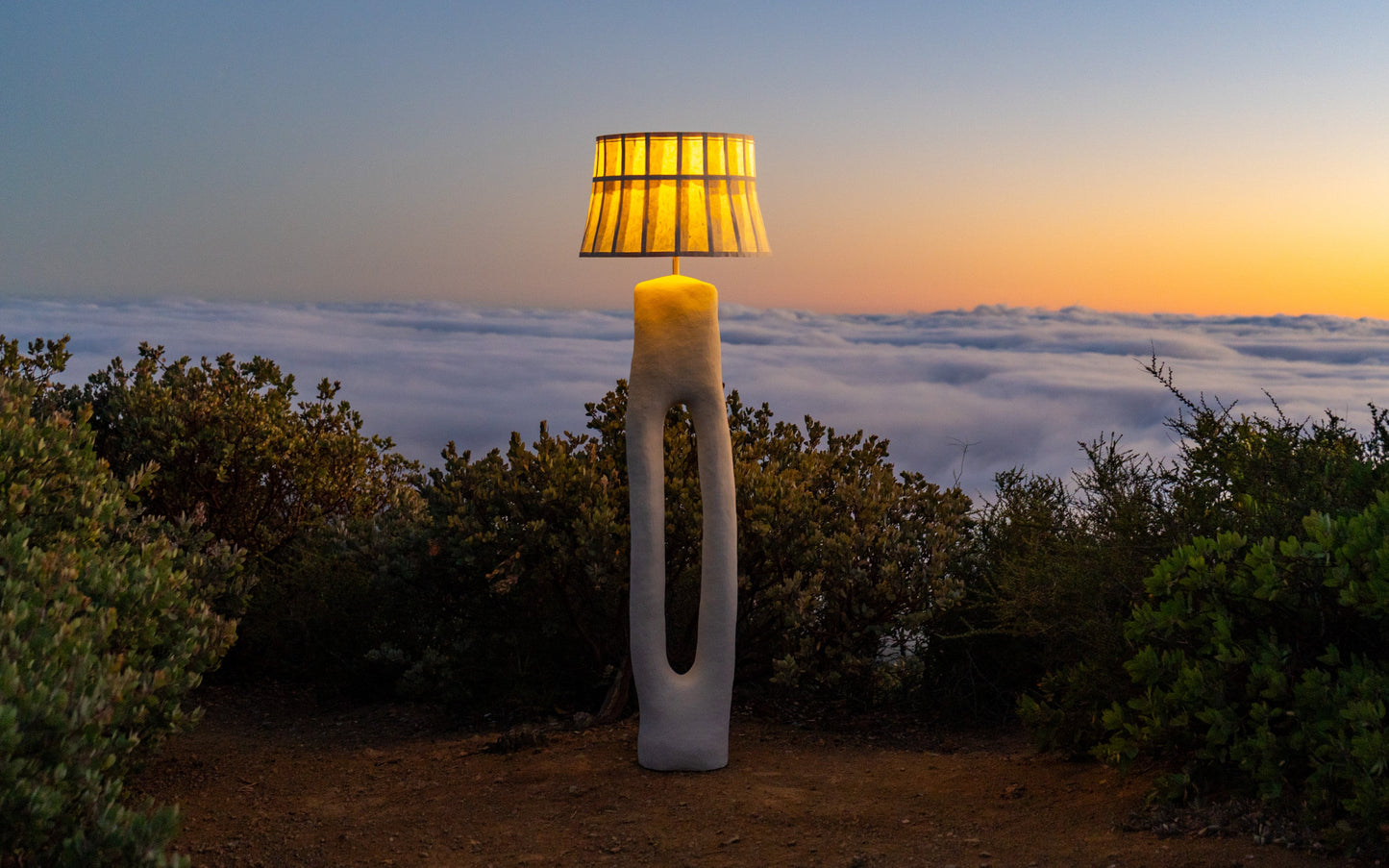 A sculptural floor lamp with a mulberry  paper shade and a white curved base stands on a dirt clearing above a sea of clouds at sunset, with shrubs in the background.