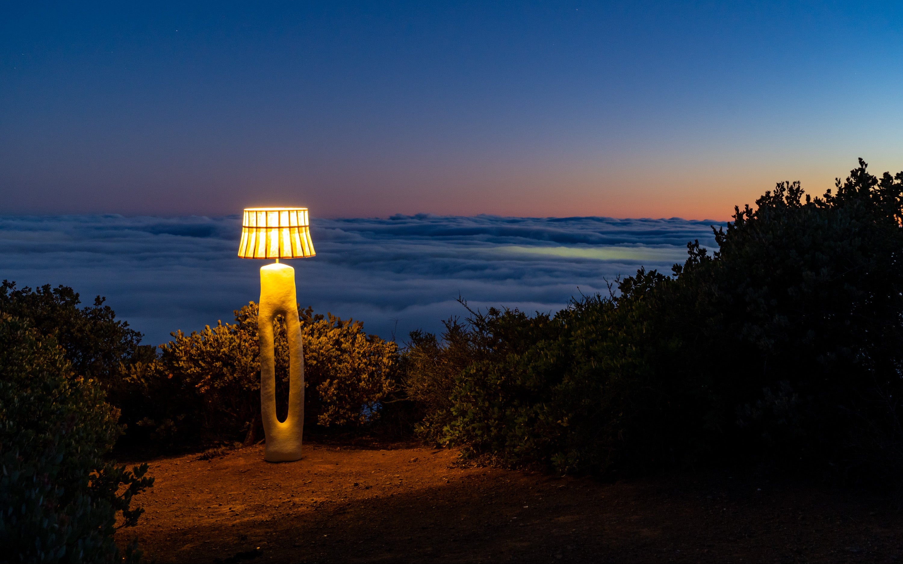 A modern outdoor floor lamp with a mulberry paper shade and sculptural base stands on a hilltop overlooking a sea of clouds at dusk, casting a warm yellow glow.