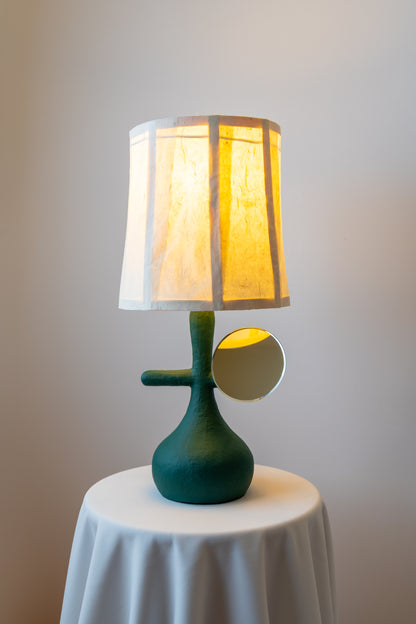 A modern table lamp with an emerald green sculptural base and a mulberry paper drum shade glowing warm yellow, displayed on a white draped table against a neutral wall.