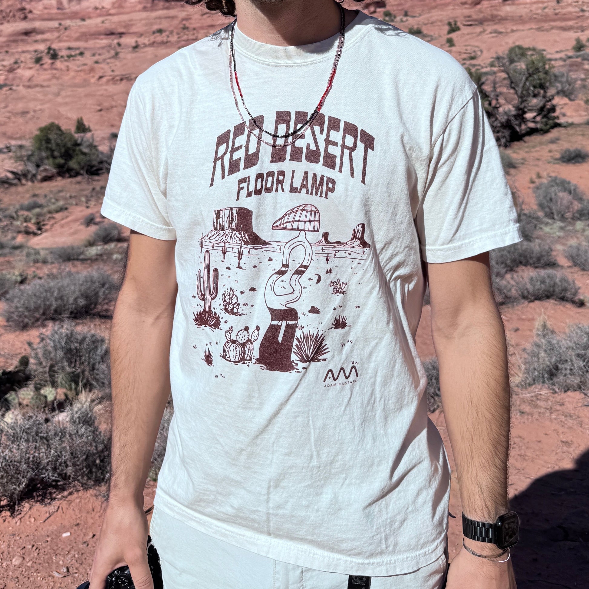 Man wearing sunglasses and a graphic t-shirt in a desert landscape with red rock formations.